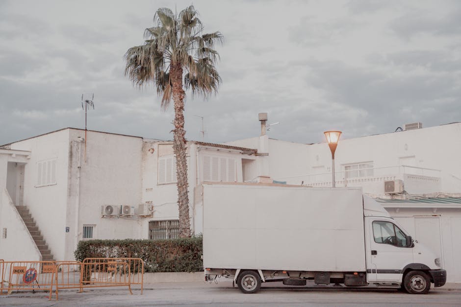 A white moving van is parked on the street outside a white residential building, part of a home relocation process. The van's side door is closed, and it appears ready for furniture transport or packi