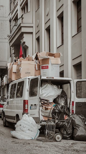 The back of a white van used for home relocation is shown parked on a city street beside a large, multi-storey building with a beige facade and several windows. The van's rear doors are open, revealing packed cardboard boxes of various sizes, some sealed with tape, and large plastic bags filled with packing materials. Cardboard boxes are stacked on top of the van, with a few positioned on the ground nearby, ready for loading or unloading. A small hand trolley is leaning against the open rear of the van. The scene captures a loading process typical of furniture transport and packing during a move, with natural daylight illuminating the area. The vehicles and packing supplies are organized to facilitate efficient house removals, aligned with services offered by Man and Van Harringay, as indicated by the context of the Harringay Green Lanes moving guide.