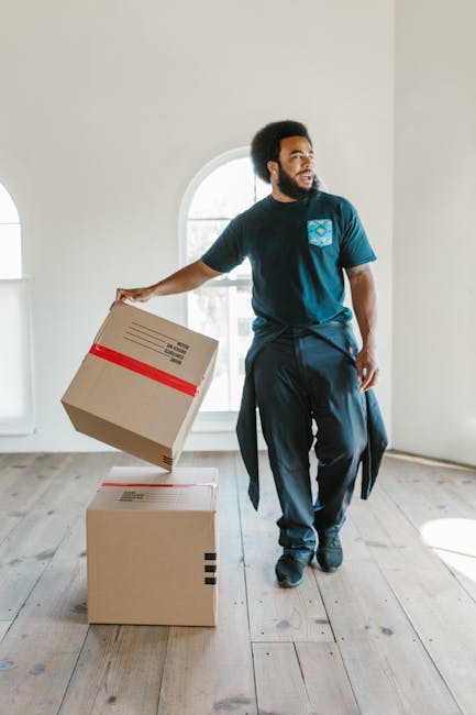 A man with dark curly hair and a beard, wearing a blue T-shirt and dark trousers, is inside a bright, spacious room with white walls and wooden flooring. He is holding a cardboard box wrapped with red