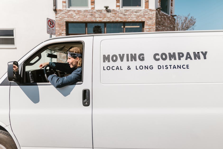 A smiling man with a full beard and curly black hair stands outdoors next to a white moving van with the company name 'Moving Company' and the words 'Local & Long Distance' printed on the side. He is 