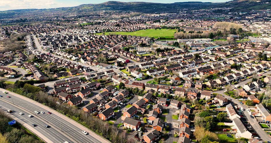 Aerial view of a residential neighbourhood in Harringay, showing rows of terraced houses, semi-detached homes, and detached properties with paved driveways, small front gardens, and rooftops made of tiles. The scene also includes a major road with moving vehicles at the bottom left, lined with trees, and a green open space in the distance. Inside a property, a moving process is visible with cardboard boxes, plastic-wrapped furniture, and wooden shelving being carried by movers wearing casual clothing. A large van, likely belonging to Man and Van Harringay, is parked nearby, with some furniture and boxes being loaded into it using a trolley and lifting straps, indicating a home relocation or furniture transport operation as part of packing and moving services. The environment is well-lit with natural daylight, and the image captures the coordination involved in house removals within the N4 postcode area, supporting efficient logistics for local property moves.