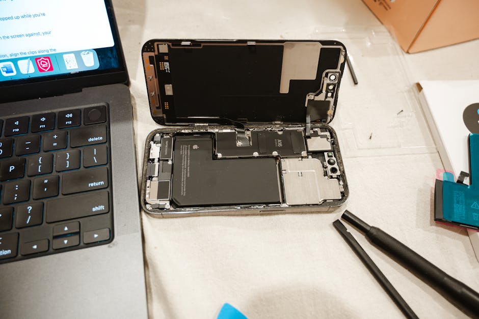 A disassembled smartphone with its screen detached and internal components, including the battery, motherboard, and connectors, visible inside the device's metal frame, situated on a white surface next to a partially visible laptop keyboard. Nearby, there are small tools such as a prying tool and a spudger, indicating a repair or refurbishment process, which is typical during packing and preparation for home relocation or furniture transport. The image depicts a technical element of packing or moving electronics as part of a professional removal service by [COMPANY_NAME], supporting smooth home moving logistics within the context of house removals and comprehensive packing and moving services on the N4 postcode area for Harringay residents.