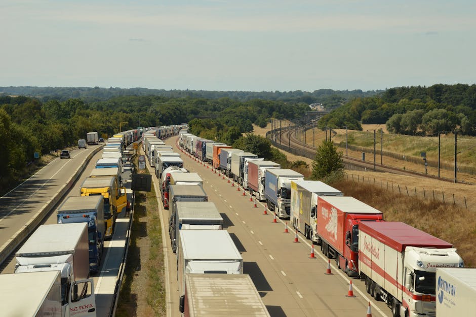 A long line of trucks and moving vans parked along a countryside road, with some vehicles positioned behind orange traffic cones to form a temporary loading zone. The trucks display various colors and branding, with some showing closed cargo areas covered by tarps or plastic wraps, indicating ongoing packing or moving operations. The scene captures a rural environment with green fields, scattered trees, and a railway track running parallel in the background. The sky is partly cloudy with diffused sunlight illuminating the scene, emphasizing the logistics involved in home relocation or furniture transport. This image relates to the moving and removals services offered by Man and Van Harringay, illustrating a possible scenario of delays or organized loading processes during a house move, aligning with the page about same day emergency removals in Harringay.