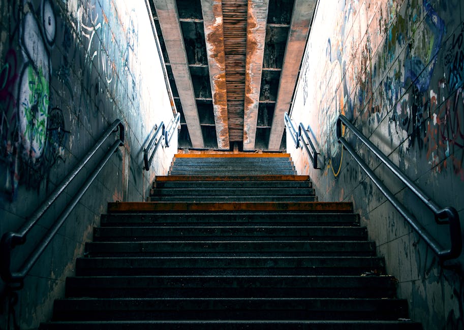 A set of dark, worn concrete stairs ascending inside an underpass or subway tunnel, with metal handrails on both sides. The stair treads show signs of rust and weathering. Graffiti covers the tiled walls on either side, with various tags and artwork in vibrant colors. Overhead, there is an exposed wooden and metal structure supporting the ceiling, which is poorly lit by ambient light filtering from above the staircase. The environment appears dingy and aged, reflecting the challenges of navigating tight or inaccessible areas during home relocation or furniture transport, consistent with the services offered by Man and Van Harringay for removals involving difficult staircases and access points.