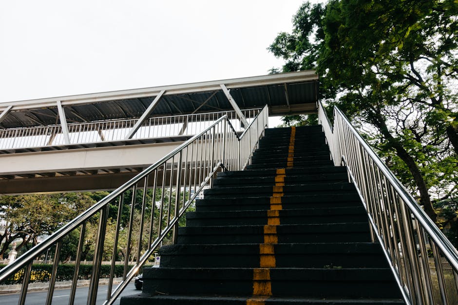 A set of dark, worn concrete stairs ascending inside an underpass or subway tunnel, with metal handrails on both sides. The stair treads show signs of rust and weathering. Graffiti covers the tiled walls on either side, with various tags and artwork in vibrant colors. Overhead, there is an exposed wooden and metal structure supporting the ceiling, which is poorly lit by ambient light filtering from above the staircase. The environment appears dingy and aged, reflecting the challenges of navigating tight or inaccessible areas during home relocation or furniture transport, consistent with the services offered by Man and Van Harringay for removals involving difficult staircases and access points.