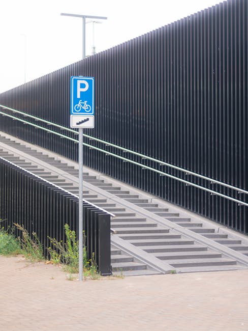 A blue parking sign indicating bicycle parking is positioned beside a sloped set of outdoor stairs with metal railings, leading up alongside a tall, dark vertical slat fence. The stairs are constructed of concrete or similar material, with visible treads and risers, situated on a paved area. The background features a minimalist, modern urban environment with a bright, clear sky overhead and a tree top visible beyond the fence. This scene is relevant to house removals and relocation logistics, demonstrating accessible entry points and the environment where furniture transport or packing and moving activities might take place, as handled by companies like Man and Van Harringay.
