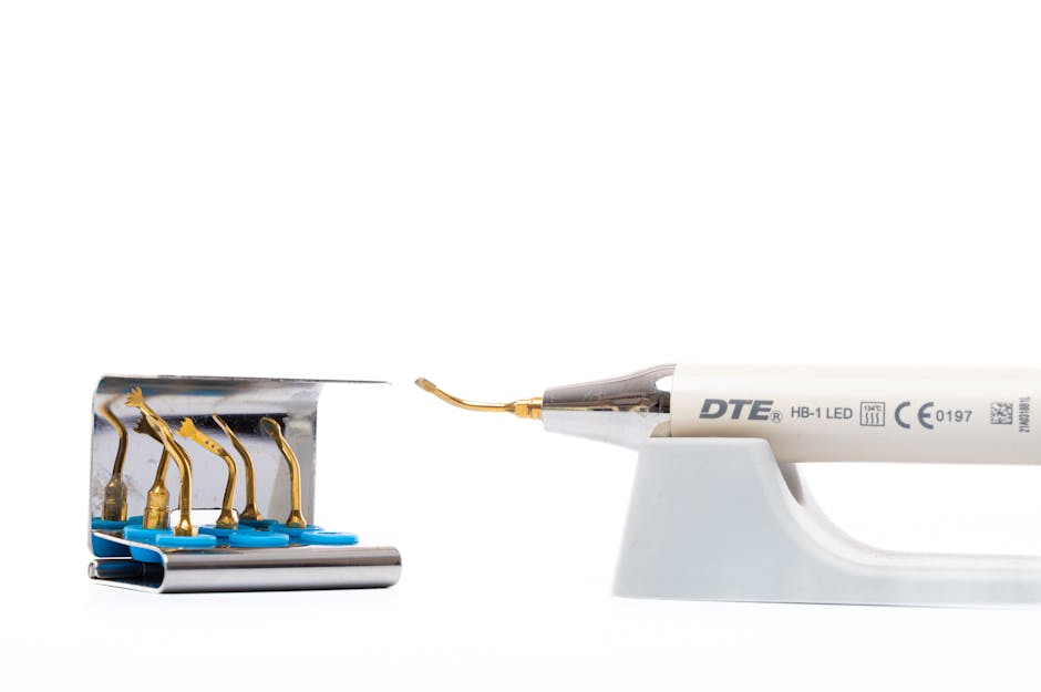 A close-up view of a dental syringe with a needle, positioned to the right, resting on a white plastic stand. To the left, there is a small metallic box containing five dental burs with gold-colored tips, each attached to blue plastic bases and arranged in a row. The background is plain white, emphasizing the medical tools, which are related to dental procedures often performed during packing and preparation phases of house removals. The image reflects the detailed and careful handling required in home relocation processes, with the tools neatly organized for easy access during the packing or setup of dental appliances in the moving logistics. Man and Van Harringay occasionally handles the careful transport of sensitive or specialized equipment, similar to the dental tools shown, as part of their removals services.